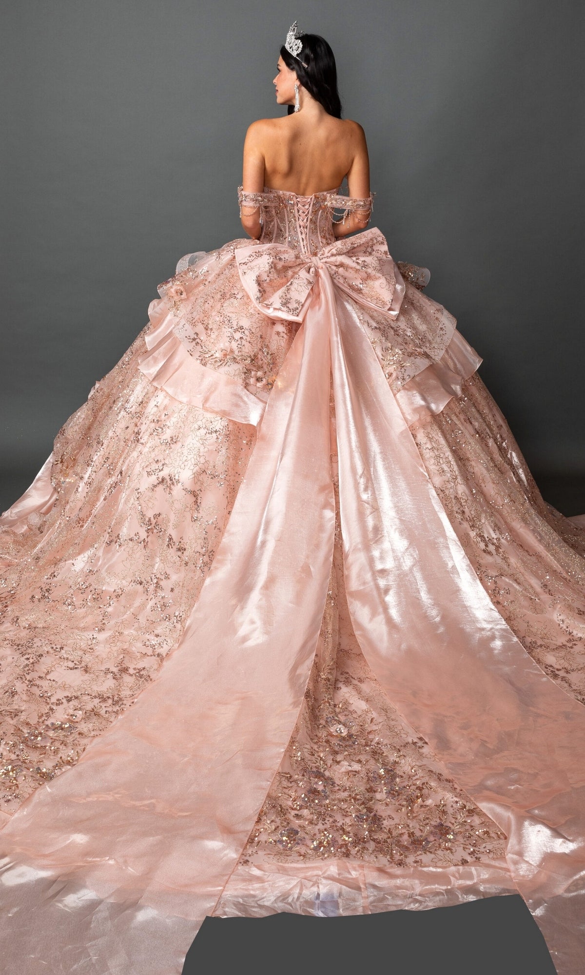 A woman poses with her back to the camera against a plain gray background, showcasing the Quinceanera Dress 5157 By Dancing Queenโan elaborate pink ball gown adorned with sequins, ruffles, 3D floral appliques, a large bow, and a dramatic long train.